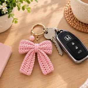 Peut inclure: Un porte-clés en métal doré avec une breloque en forme de nœud crocheté rose clair et une perle rose ronde. Le porte-clés est attaché à une clé en métal argenté et à un boîtier de clé de voiture Honda noir avec quatre boutons de verrouillage.