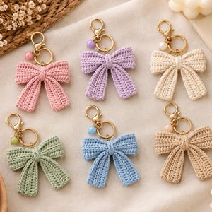 May include: Six crochet bow keychains in pink, lavender, cream, sage green, light blue, and tan. Each bow is attached to a gold-tone metal lobster clasp and keyring, accented with a single small round bead in a coordinating pastel colour.