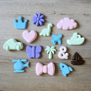 May include: A collection of fifteen small, matte-finish silicone shapes in pastel colours. The items include a blue crown, purple flower, pink cloud, light green dinosaur, pink heart, blue aeroplane, light green boat, light green car, purple butterfly, light green palm tree, pink ampersand, brown sailboat, blue helicopter, pink bow, and a blue rocket ship.