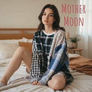 May include: A long-sleeve crewneck sweatshirt featuring a patchwork design of various plaid and checkered patterns in black, white, blue, and grey. The text MOTHER MOON is printed in a reddish-pink font in the upper right corner of the image.