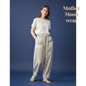 以下が含まれることがあります： 無地のクリーム色の半袖Tシャツと、ベージュと白のチェック柄のゆったりとしたパンツの組み合わせ。パンツは伸縮性のあるドローストリングウエストで、パッチワークのディテールが施されたフロントポケットが2つ付いています。Mother Moon wearというテキストが表示されています。