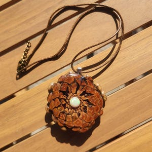 May include: A brown pinecone pendant necklace featuring a central iridescent white cabochon stone. The pendant is attached to a thin brown cord with a metallic clasp and an adjustable chain link extension, creating a natural and textured appearance.