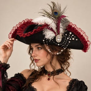 May include: A black tricorn hat featuring a deep red ruffled lace trim, white and peacock feathers, and a central oval rhinestone brooch. The hat is adorned with delicate strands of white pearls and scattered clear crystals across the side.