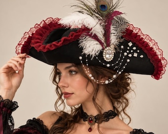 May include: A black tricorn hat featuring a deep red ruffled lace trim, white and peacock feathers, and a central oval rhinestone brooch. The hat is adorned with delicate strands of white pearls and scattered square crystals across the side.