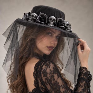 May include: A black hat featuring a wide brim and a flat crown, adorned with a band of silver-toned skull figures interspersed with black fabric roses. A sheer black tulle veil hangs from the brim, draping over the front of the hat.