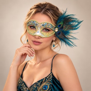 May include: A masquerade mask featuring a white base with gold filigree, blue accents, and clear rhinestones. The mask is adorned with a large cluster of teal and dark blue peacock feathers on the right side, accented by a single round clear crystal.