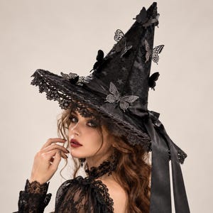 May include: A black pointed hat featuring a wide brim with intricate lace trim. The hat is decorated with multiple three-dimensional butterfly embellishments and a satin ribbon band. The fabric has a subtle star pattern throughout the surface.