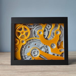 May include: A rectangular black frame containing a layered mechanical display of interlocking gears and cogs. The composition features a mix of bright yellow and light grey plastic components, showcasing various circular gear sizes and curved arms.