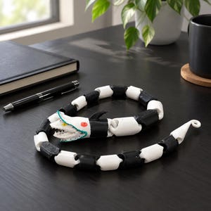 May include: A segmented, articulated snake figurine featuring a repeating pattern of alternating black and white cylindrical links. The head is white with a teal trim, sharp teeth, a yellow eye, and a red eye, ending in a curled tail tip.