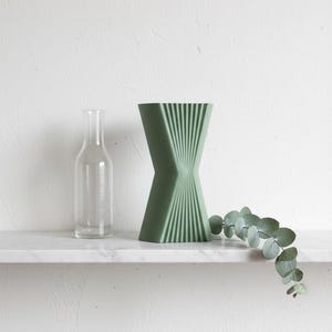 May include: A sage green vase with a pinched centre and vertical ribbed texture, accompanied by a clear glass bottle and a single sprig of eucalyptus leaves. The vase features a flared top and base with a distinct geometric hourglass shape.