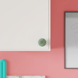 May include: A round cabinet knob with a textured, sage green surface featuring a cracked, organic pattern. The knob is mounted on a flat, white cabinet door panel, providing a distinct contrast against the smooth, solid white surface of the door.