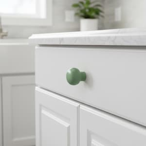 May include: A round, sage green cabinet knob with a smooth, matte finish attached to a white, flat-panel drawer front. The knob features a simple, spherical shape that protrudes from the center of the drawer, providing a clean, functional accent.