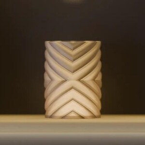 May include: A cylindrical lamp with a matte beige finish featuring a repeating geometric chevron pattern that wraps around the entire exterior. The structure emits a soft, warm glow from within, highlighting the textured ridges and valleys.