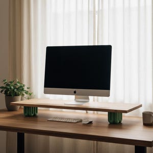 May include: A light-coloured wooden monitor stand featuring a flat rectangular top surface supported by two sage green, vertically ribbed, cylindrical legs. The stand is positioned on a wooden desk, holding a silver computer monitor with a black screen.