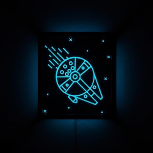 May include: A square wall-mounted light box featuring a glowing cyan neon-style outline of a circular spaceship with a trailing motion effect. The design includes small cross-shaped stars against a solid black background, emitting a soft blue light.