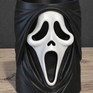 May include: A cylindrical black container featuring a white, elongated, ghost-like mask with hollow eyes and an open mouth. The mask is integrated into the front of the black vessel, which has a smooth, matte finish and a draped, fabric-like texture.