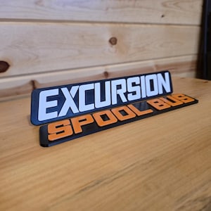 May include: A two-part rectangular sign featuring raised lettering. The top section is a black plaque with white text reading EXCURSION. The bottom section is a black plaque with orange text reading SPOOL BUS. Both pieces have a matte finish.