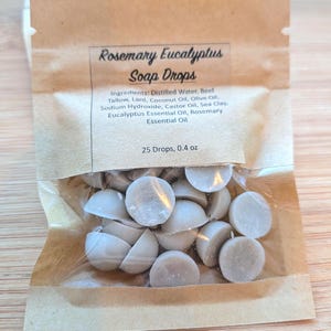 May include: A brown paper pouch containing 25 gray, semi-spherical soap drops. The label reads Rosemary Eucalyptus Soap Drops, listing ingredients including beef tallow, lard, coconut oil, olive oil, and essential oils, totaling 0.4 oz.