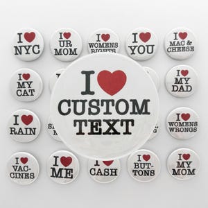 May include: A collection of circular white pin-back badges featuring black text and a red heart symbol. The central badge displays the text I HEART CUSTOM TEXT. Surrounding badges feature various phrases including I HEART NYC, I HEART MY CAT, I HEART MY MOM, I HEART MY DAD, I HEART ME, I HEART CASH, I HEART BUTTONS, and I HEART VACCINES.