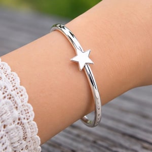 May include: A polished silver-tone bangle bracelet featuring a single five-pointed star charm centred on the band. The bracelet has a smooth, reflective metallic finish and a simple, open-ended design that curves around the wrist.