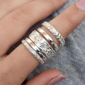 May include: A stack of five silver-toned rings worn on a finger. The rings feature varied textures including a braided pattern, a smooth polished band, a floral engraved design, a plain band, and a band with a fine, intricate etched surface texture.