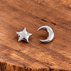 May include: A pair of silver-toned stud earrings featuring a five-pointed star shape and a crescent moon shape. Both earrings have a smooth, polished, reflective metallic finish and are presented as individual pieces side by side.