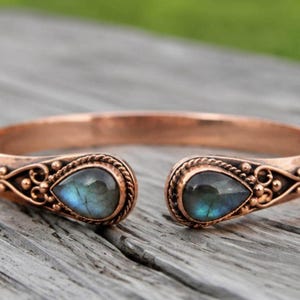May include: A copper cuff bracelet featuring an open design with two teardrop-shaped labradorite gemstones at the ends. The metal band displays intricate wire-wrapped detailing and small decorative spheres surrounding each stone setting.