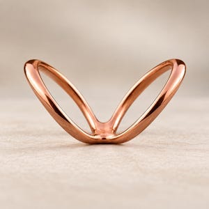 May include: A polished rose gold ring featuring a V-shaped design with two smooth, curved bands that meet at a single point. The metal has a reflective, metallic finish and a continuous, open structure that creates a symmetrical, wing-like appearance.