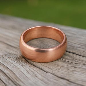 May include: A solid copper ring with a smooth, polished finish and a rounded, domed profile. The band features a warm, metallic reddish-brown hue with a consistent, reflective surface that catches the light, showcasing a simple, circular design.