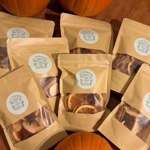 Op de afbeelding: Zes hersluitbare, beige stazakken met gedroogde sinaasappelschijfjes, kaneelstokjes en steranijs. Elke zak is voorzien van een rond wit etiket met de tekst Cozy Fall Simmerpot en een lijst met ingrediënten.