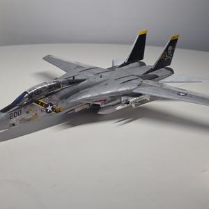 Może przedstawiać: Szary model myśliwca Grumman F-14 Tomcat z numerem 200 na nosie, przezroczystą owiewką kabiny oraz czarnymi statecznikami pionowymi z emblematem czaszki i skrzyżowanych piszczeli. Samolot jest wyposażony w pociski rakietowe.