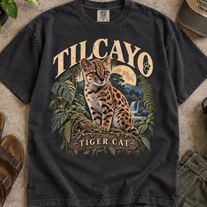 Peut inclure: Un t-shirt gris anthracite à manches courtes avec une impression graphique représentant un chat tigre tacheté assis sur une branche. Le motif inclut un feuillage tropical, une cascade, une pleine lune et le texte TILCAYO et TIGER CAT en beige.