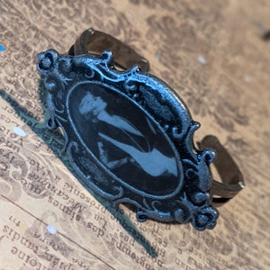 May include: A silver ring with an oval-shaped cameo featuring a black and white image of a woman. The ring has a detailed ornate frame.