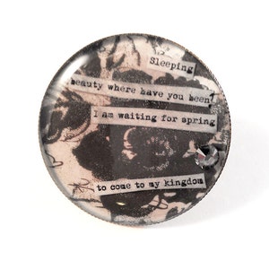 May include: A round, glass, decorative paperweight with a black and white floral design and a quote that reads "Sleeping beauty where have you been? I am waiting for spring to come to my kingdom".