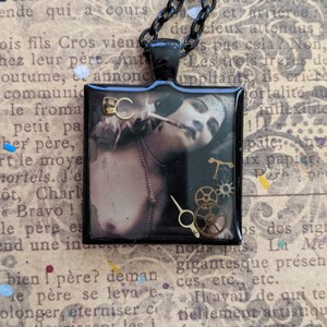 May include: A black necklace with a square pendant featuring a woman with a black eye mask and a steampunk-inspired design. The pendant has a gold-colored gear and a gold-colored T-shaped piece.