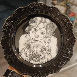 Puede incluir: Un plato decorativo negro con un borde festoneado y en relieve que presenta una ilustración de líneas blancas en el centro. La obra muestra a una persona con cabello hasta los hombros abrazando a un gato esponjoso, en un estilo de boceto detallado.