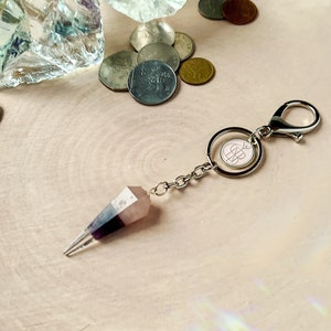 May include: A silver keyring with a pink and purple crystal point charm. The charm is attached to a silver chain and a silver ring with a pink circle containing a monogram.