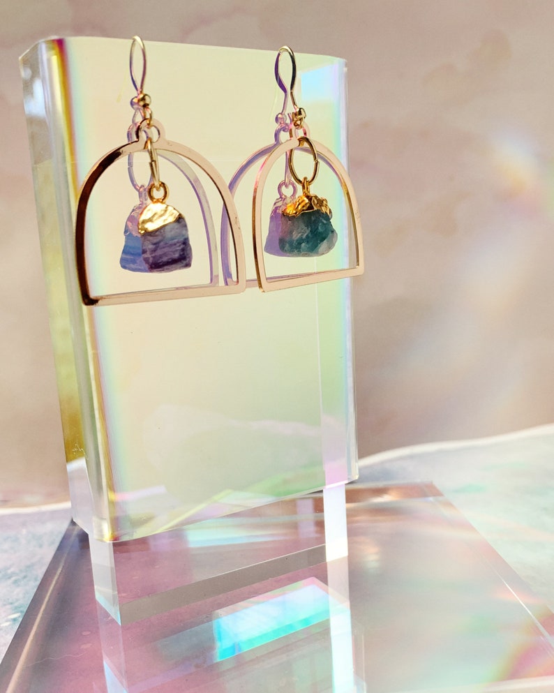 Pode incluir: Brincos de argola dourados com uma gema azul e roxa de forma irregular pendurada. A gema &eacute; adornada com folha de ouro.