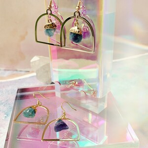 Pode incluir: Brincos de argola de ouro com uma pedra de cristal azul-esverdeada e roxa pendurada em cada argola.