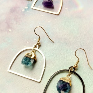 Pode incluir: Um par de brincos de ouro e prata em forma de arco com uma pedra de cristal pendurada. Os brincos est&atilde;o dispon&iacute;veis em duas cores: azul e roxo.