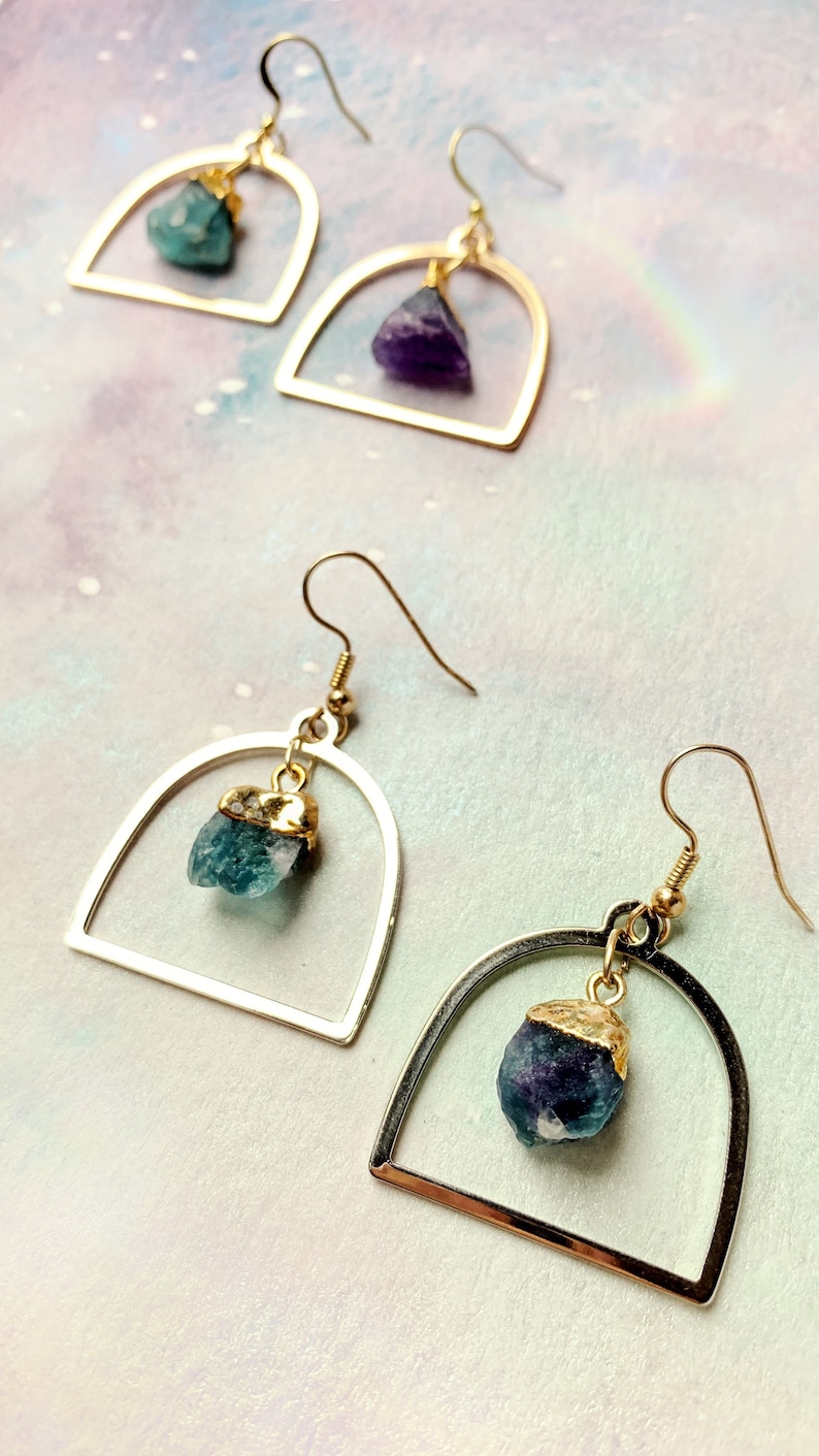 Pode incluir: Um par de brincos de ouro e prata em forma de arco com uma pedra de cristal pendurada. Os brincos est&atilde;o dispon&iacute;veis em duas cores: azul e roxo.