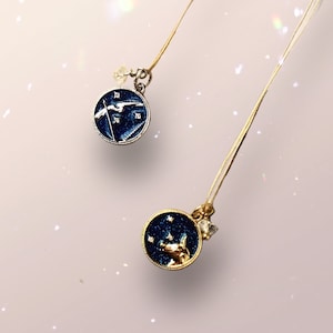May include: Two gold necklaces with blue and gold pendants. The pendants are round and have a starry night design. One pendant features a silver bird, the other features a gold animal.