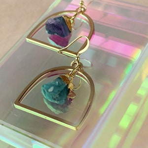 Pode incluir: Brincos de argola dourados com uma gema azul-esverdeada irregular pendurada. Os brincos est&atilde;o sobre uma superf&iacute;cie branca iridescente.