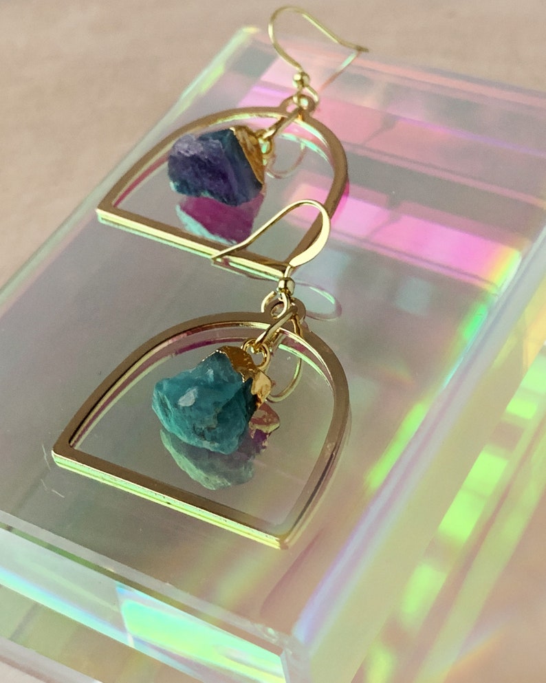 Pode incluir: Brincos de argola dourados com uma gema azul-esverdeada irregular pendurada. Os brincos est&atilde;o sobre uma superf&iacute;cie branca iridescente.