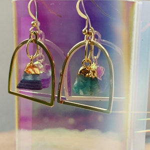 Pode incluir: Brincos de argola dourados com uma pedra de cristal verde e roxa pendurada. Os brincos s&atilde;o exibidos em um fundo iridescente.