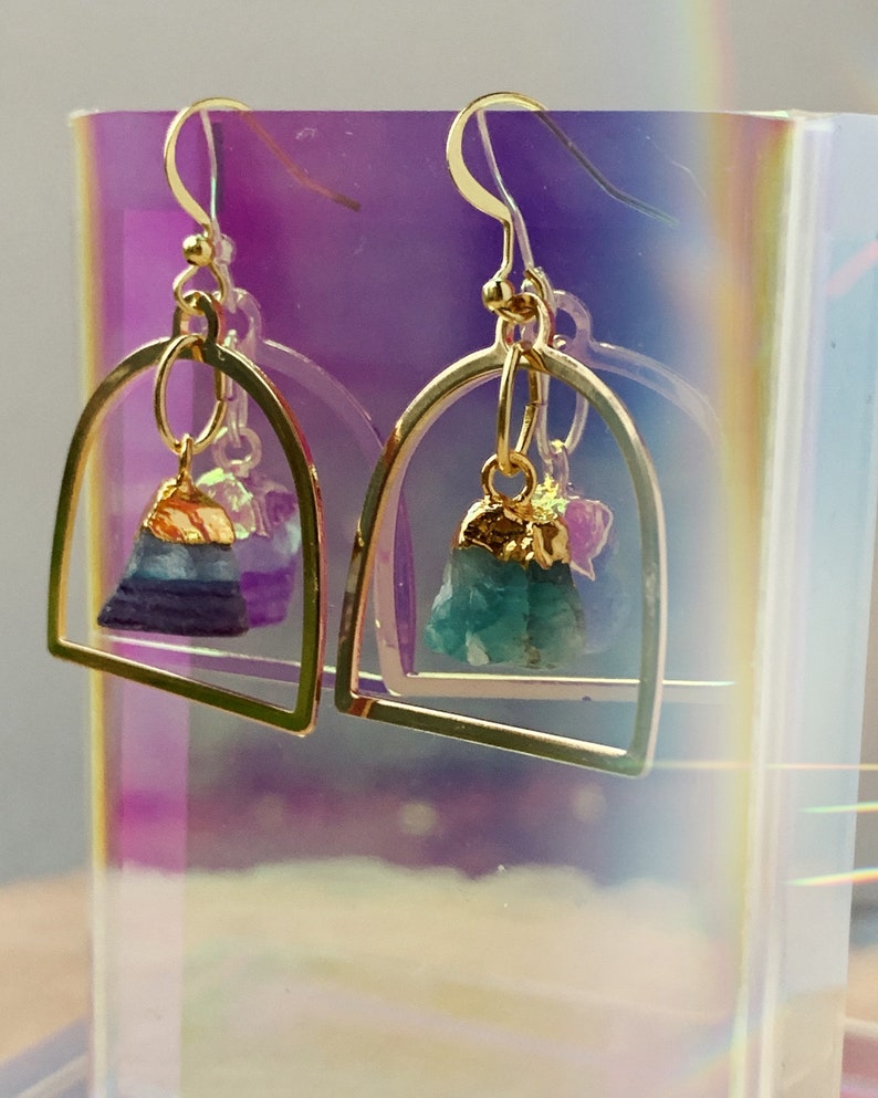 Pode incluir: Brincos de argola dourados com uma pedra de cristal verde e roxa pendurada. Os brincos s&atilde;o exibidos em um fundo iridescente.