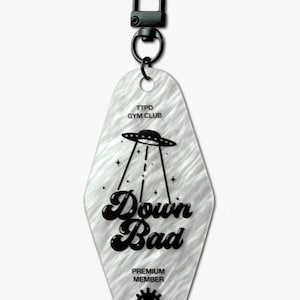 Pode incluir: Um chaveiro branco texturizado com um fecho de metal preto. O chaveiro apresenta o texto "TTPD GYM CLUB" e "Down Bad" com uma ilustração de OVNI. A parte inferior diz "PREMIUM MEMBER" com um olho estilizado.