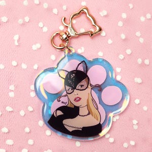 Peut inclure: Un porte-clés transparent en forme de patte avec un fermoir doré. Le porte-clés présente une illustration de dessin animé d'une femme portant un masque de chat, avec des accents roses et bleus. Le fond est un tissu rose à pois blancs.