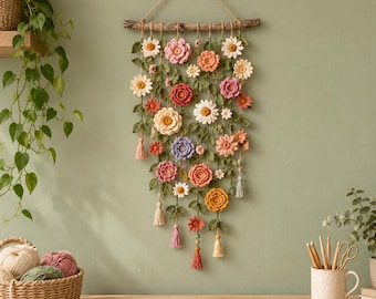 Puede incluir: Un tapiz de pared que presenta una rama de madera horizontal con múltiples hilos de flores de ganchillo en tonos rosa, naranja, amarillo, blanco y morado, acentuado con hojas de ganchillo verdes y pequeñas borlas colgantes.