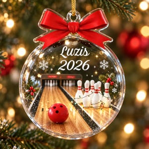 May include: A clear, flat, circular Christmas ornament featuring a red bow at the top. The center displays a bowling alley scene with a red bowling ball and white pins. The text "Luzis" and "2026" are printed in white, surrounded by snowflake icons.